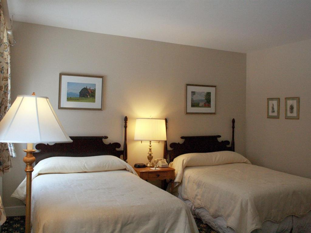 A room with two double beds and white bedding with wooden bed frames sit in the middle of the room. An antique light on a bedside table is in between the beds.